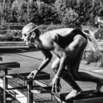 Dive Preparation. - Black and white side view full body sporty swimmer in swimming suit and goggles standing on block in track start position preparing to dive in outside pool