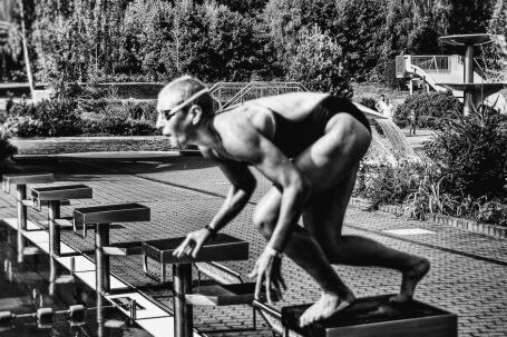 Dive Preparation. - Black and white side view full body sporty swimmer in swimming suit and goggles standing on block in track start position preparing to dive in outside pool