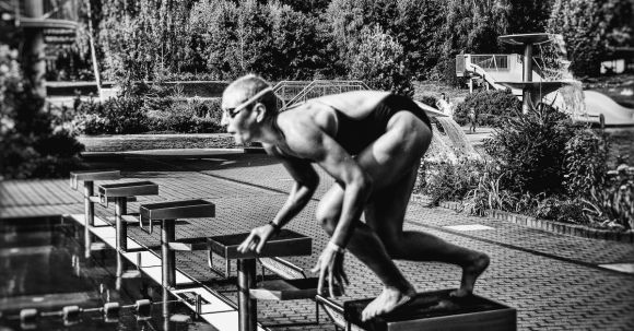 Dive Preparation. - Black and white side view full body sporty swimmer in swimming suit and goggles standing on block in track start position preparing to dive in outside pool