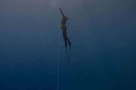 Wetsuit - Side view of anonymous diver in wetsuit and flippers swimming up rope under transparent crystal blue seawater close to surface