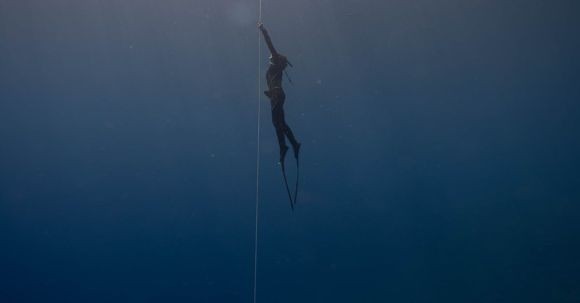 Wetsuit - Side view of anonymous diver in wetsuit and flippers swimming up rope under transparent crystal blue seawater close to surface