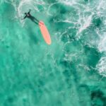 Wetsuit - Drone view of faceless surfer in wetsuit swimming near longboard in vibrant turquoise lagoon in sunlight