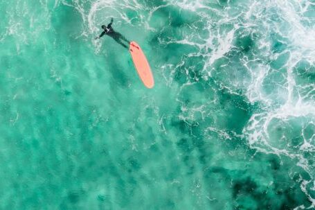 Wetsuit - Drone view of faceless surfer in wetsuit swimming near longboard in vibrant turquoise lagoon in sunlight