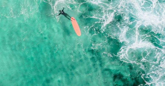 Wetsuit - Drone view of faceless surfer in wetsuit swimming near longboard in vibrant turquoise lagoon in sunlight