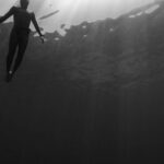 Wetsuit - Black and white of from below of scuba diver swimming in clear water in swimsuit under rays of light