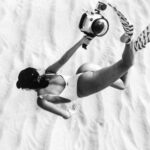 Underwater Camera - Black and white faceless slim female wearing swimsuit and flippers snorkeling above sandy sea bottom in transparent waters and carrying camera in waterproof cover