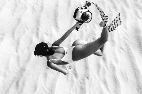 Underwater Camera - Black and white faceless slim female wearing swimsuit and flippers snorkeling above sandy sea bottom in transparent waters and carrying camera in waterproof cover