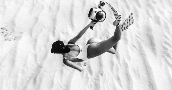 Underwater Camera - Black and white faceless slim female wearing swimsuit and flippers snorkeling above sandy sea bottom in transparent waters and carrying camera in waterproof cover